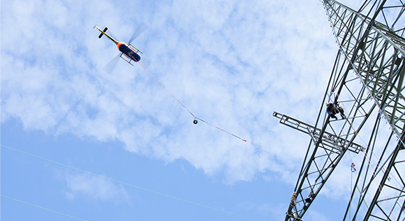 Hubschrauber schwebt über Strommast, an dem Wartungsarbeiten durchgeführt werden.