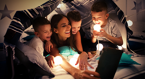 Eine junge Mutter mit ihren drei kleinen Söhnen spielt in einer selbstgebauten Höhle mit leuchtenden Energiesparlampen